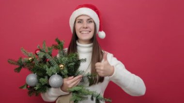 Güzel, gülümseyen genç bir bayan beyaz süveter giyer ve Noel şapkası demetinde ladin dalları kırmızı stüdyo arka plan duvarında izole edilmiş bir jest gibi başparmağını kaldırır. Mutlu yıllar tatil konsepti