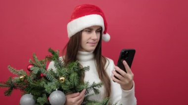 Mutlu genç kızın portresi beyaz süveter giyer ve Noel şapkası demetinde ladin dalları kırmızı arka planda izole edilmiş şekilde cep telefonu kullanır. Yeni yıl tatili konsepti