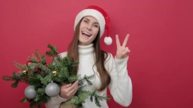 Noel Baba şapkalı genç ve mutlu bir kadının portresi elinde ladin demeti tutarak stüdyoda kırmızı renkli arka plan duvarı üzerinde izole edilmiş bir şekilde zafer işareti gösteriyor. Mutlu yıllar 2023 tatil konsepti