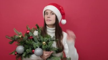 Meraklı genç kadın beyaz kazak giyiyor ve elinde ladin demeti olan Noel Baba şapkası var. Kulak misafiri olmaya çalış. Stüdyodaki kırmızı arka plan duvarında izole bir şekilde poz veriyorsun.