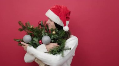Mutlu bir portre genç Noel Baba kadın beyaz kazak giyiyor ve Noel şapkası tutuyor. Stüdyonun kırmızı arka plan duvarında izole edilmiş ladin dalları buketine bakın. Mutlu Yıllar Kutlama Konsepti
