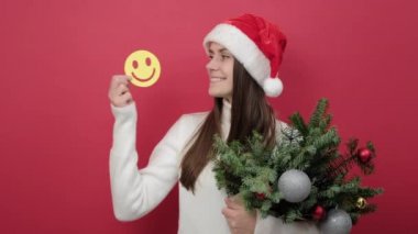 Noel Baba şapkalı mutlu genç kadının portresi ladin dalları buketi ve sarı komik yüz ifadeleri ile stüdyoda kırmızı arka planda izole edilmiş poz veriyor. Noel ve Yeni Yıl tatili konsepti