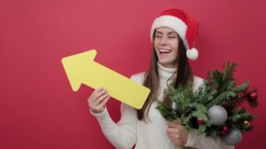 20 'li yaşlarda genç bir kadının portresi beyaz süveter ve kırmızı stüdyo arka planında izole edilmiş ladin dalları ve sarı oklardan oluşan bir buket Noel Baba şapkası giyer. Mutlu yıllar 2023 tatil konsepti