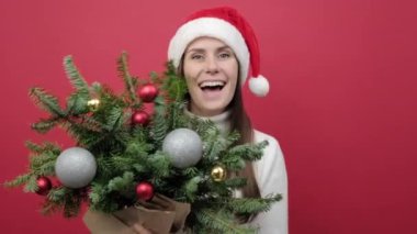 Mutlu genç Noel Baba kadın portresi beyaz süveter giyer ve Noel şapkası askısı takar ladin dallarının arkasına saklanır, kırmızı stüdyo arka plan duvarında izole edilmiş poz verir. Mutlu Yıllar Kutlama Konsepti