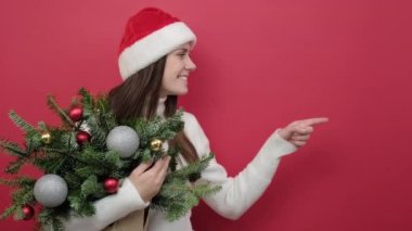 Şaşırmış genç Noel Baba kadının portresi Noel şapkası takıyor ladin demeti çalışma alanındaki nokta fotokopi alanı, stüdyo odasındaki kırmızı arkaplan duvarının üzerinde izole edilmiş şekilde duruyor.