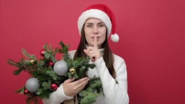 Gizli esmer kız portresi 20 'li yaşlarda beyaz süveter giyer ve Noel Baba şapkalı ladin demeti der ki sessiz ol dudaklarında parmak, dudaklarında jest, kırmızı stüdyo arka planında izole edilmiş.