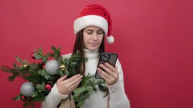 Neşeli genç esmer kadının portresi beyaz kazak giyer ve Noel Baba şapkalı ladin demeti kırmızı stüdyo arka planında izole cep telefonu kullanır. Mutlu yıllar 2023 tatil konsepti