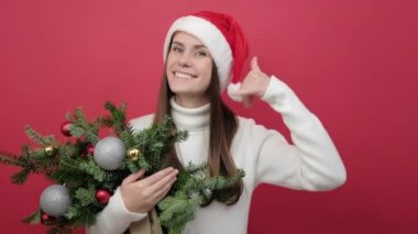 20 'li yaşlarda gülümseyen genç bir kadının portresi beyaz süveter giyer ve Noel Baba şapkası demet ladin dallarını tutar.