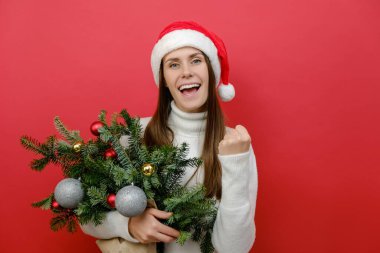 Noel Baba şapkalı neşeli beyaz kadın elinde ladin dalları buketi tutarken kazanan jestler yapıyor yumruklarını sıkarak evet diyor, kırmızı arka planda yalnız poz veriyor. Noel konsepti