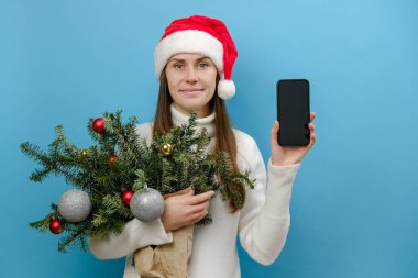 Genç esmer kadının portresi beyaz süveter giyer ve Noel şapkası tutacağı demet ladin dalları atölyenin mavi arka planındaki boş ekran çalışma alanı olan cep telefonu kullanır.