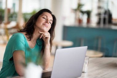 Düşünceli, düşünceli, kumandalı, koyu renk saçlı bir kadın bir dizüstü bilgisayarın ya da defterin önünde çalışma masasında oturuyor. Modern, parlak oturma odasında, bir sürü penceresi olan çalışma masasında.