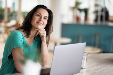 Düşünceli, düşünceli, kumandalı, koyu renk saçlı bir kadın bir dizüstü bilgisayarın ya da defterin önünde çalışma masasında oturuyor. Modern, parlak oturma odasında, bir sürü penceresi olan çalışma masasında.