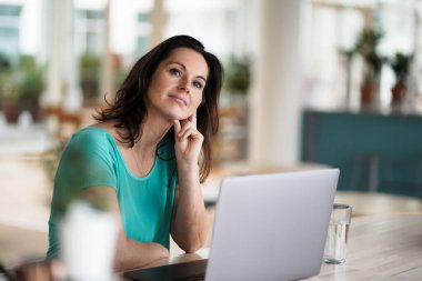 Düşünceli, düşünceli, kumandalı, koyu renk saçlı bir kadın bir dizüstü bilgisayarın ya da defterin önünde çalışma masasında oturuyor. Modern, parlak oturma odasında, bir sürü penceresi olan çalışma masasında.