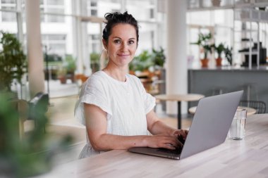 Mutlu gülümseyen, uzaktan kumandalı, koyu renk saçlı, neşeli, dizüstü bilgisayarlı, ya da günlük kıyafetli modern oturma odasındaki çalışma masasında oturmuş video sohbeti yapan mutlu bir kadın.