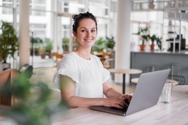 Mutlu gülümseyen, uzaktan kumandalı, koyu renk saçlı, neşeli, dizüstü bilgisayarlı, ya da günlük kıyafetli modern oturma odasındaki çalışma masasında oturmuş video sohbeti yapan mutlu bir kadın.