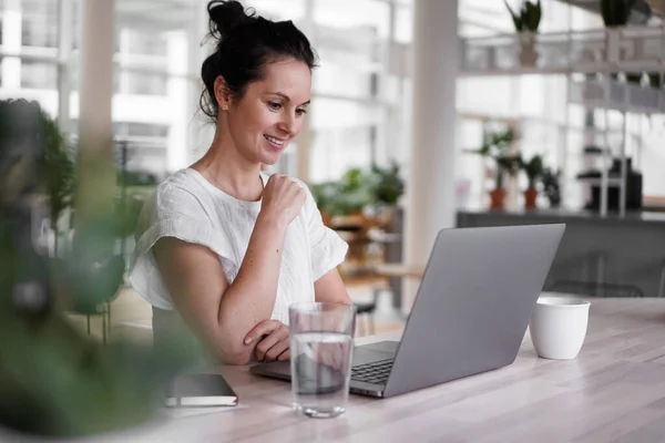 happy smiling  successful remote working dark haired woman cheerful infront of laptop or notebook in casual outfit sitting on work desk in her modern loft living room home office having a video chat