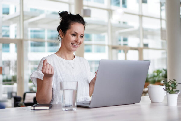 счастливая улыбающаяся удалённая работающая темноволосая женщина весёлая перед ноутбуком или ноутбуком в повседневной одежде сидящая на рабочем столе в своём современном лофт-гостиной домашнем офисе с видеочатом