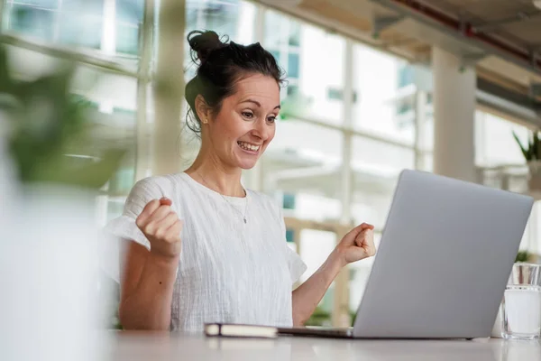 happy smiling  successful remote working dark haired woman cheerful infront of laptop or notebook in casual outfit sitting on work desk in her modern loft living room home office having a video chat