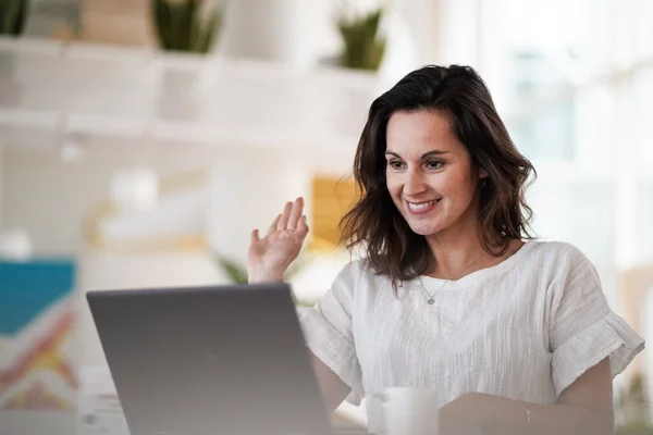 smiling remote working woman chatting and waving hand infront of a laptop or notebook during a video conference on her work desk in her modern bright living room home office