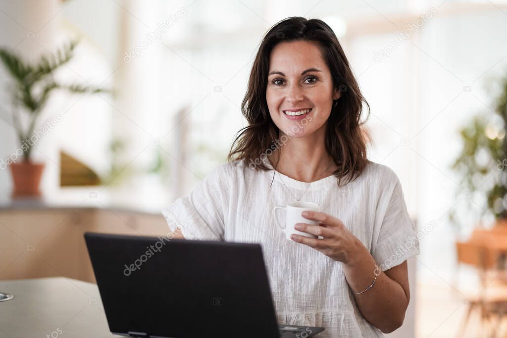 happy smiling remote working woman drinking coffee or espresso infront of a laptop or notebook on her work desk in her modern airy bright living room home office 