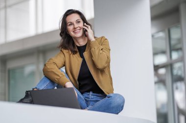 Gülümseyen, uzaktan kumandalı, çalışan bir bayan laptop ya da dizüstü bilgisayarın önünde arıyor ve sohbet ediyor. Üzerinde cep telefonu, modern bir otel lobisinde beyaz bir zemin üzerinde.
