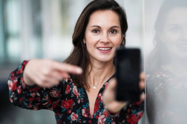 Mutlu gülümseyen şık iş kadını kırmızı çiçekli, iyimser ve neşeli bir elbise içinde cep telefonunu kameraya göstererek ön tarafı işaret ediyor.