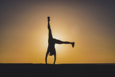 Spor yapan atletik bir kadının silüeti gün batımında plaj kaldırımında takla atıp amuda kalkıyor. 