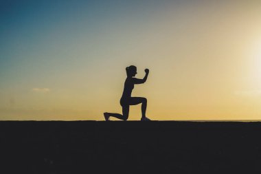 Başarılı, güçlü ve sportif bir kadının silüeti gün batımında veya gün doğumunda sahilde yumruklarıyla şampiyon pozu veriyor. 