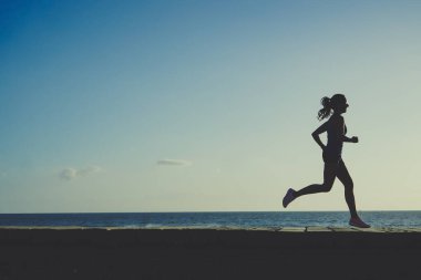 Gün batımında sahil kenarında koşan atletik bir kadının silüeti. 
