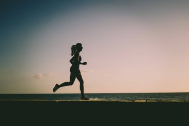Gün batımında sahil kenarında koşan atletik bir kadının silüeti. 