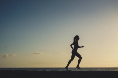 Gün batımında sahil kenarında koşan atletik bir kadının silüeti. 