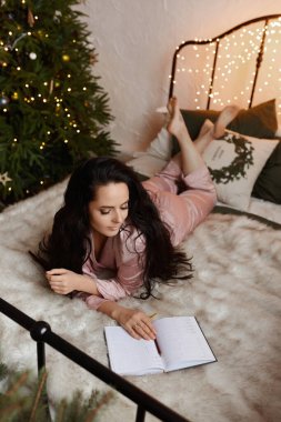 Hoş, esmer, rahat bir uyku elbisesi içinde genç bir kadın. Yatakta oturuyor ve yılbaşı için Noel ağacının yanındaki deftere yeni yıl planları yazıyor.