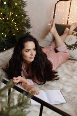 Hoş, esmer, rahat bir uyku elbisesi içinde genç bir kadın. Yatakta oturuyor ve yılbaşı için Noel ağacının yanındaki deftere yeni yıl planları yazıyor.