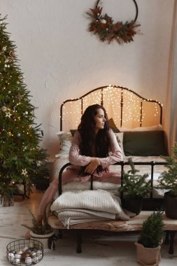 Güzel, esmer, rahat pijamalı bir kadın Noel ağacının yanındaki yatakta oturuyor.