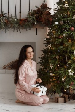 Mutlu, güler yüzlü bir kadın elinde hediye kutusuyla şık pijama giyiyor ve evinde Noel ağacının yanında poz veriyor.