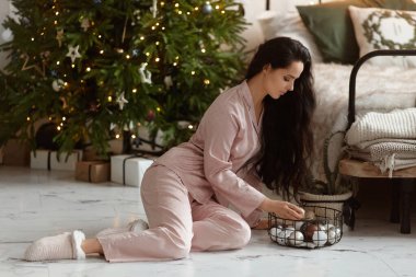 Koyu renk saçlı, elinde Noel süsleri olan ve Noel ağacının yanında poz veren son moda pijamalar giyen genç bir kadın.