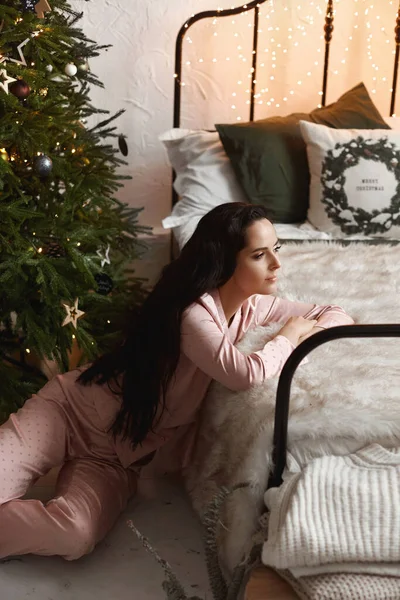 Nice ciemnowłosa dziewczyna ubrana w przytulne piżamy siedzi na podłodze w pokoju obok Nowego Roku drzewa i prezenty — Zdjęcie stockowe