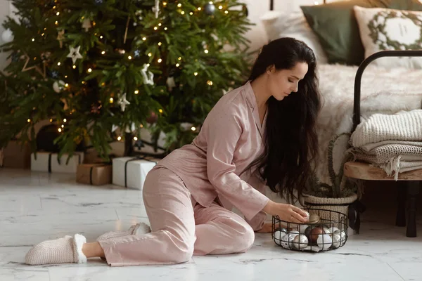 Koyu renk saçlı, elinde Noel süsleri olan ve Noel ağacının yanında poz veren son moda pijamalar giyen genç bir kadın.