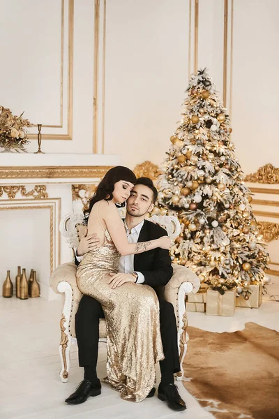 Sexy pareja apasionada celebrando la Navidad juntos. Mujer atractiva en vestido de noche de oro y hombre barbudo guapo en un traje posando en un interior con luces festivas de año nuevo en el fondo —  Fotos de Stock