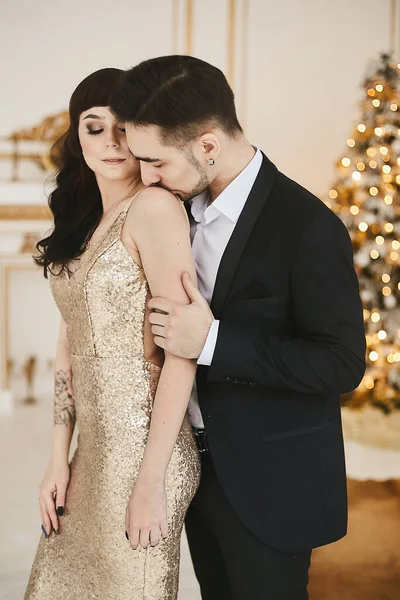 Sexy casal apaixonado celebrando o Natal juntos. Mulher atraente em vestido de noite de ouro e homem barbudo bonito em um terno posando em um interior com luzes festivas de Ano Novo no fundo — Fotografia de Stock