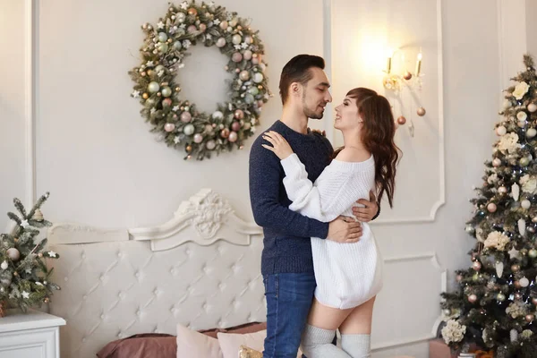 一对甜蜜的年轻夫妇在一棵装饰过的圣诞树旁的卧室里拥抱在床上 — 图库照片