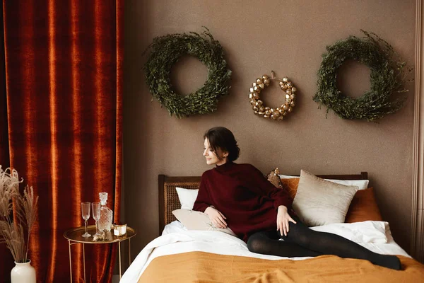 Gorgeous young woman in knitted dress sits on the bed in the interior decorated for Christmas — Stock Photo, Image