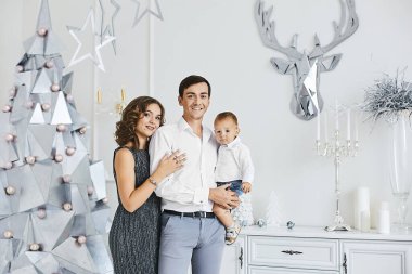Genç ebeveynler ve küçük oğulları yeni yılı ve Noel tatilini kutluyorlar. Genç bir kadın ve yakışıklı bir adam Noel için dekore edilmiş şirin küçük oğullarıyla poz veriyorlar.