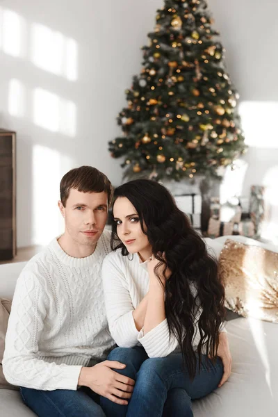 Belle fille modèle avec de longs cheveux noirs et son beau petit ami assis sur le canapé avec arbre de Noël en arrière-plan — Photo