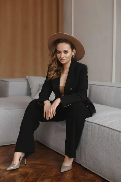 Молодая женщина с идеальным макияжем в модных шляпных позах в интерьере. Модель девушки в модном наряде сидит на диване — стоковое фото