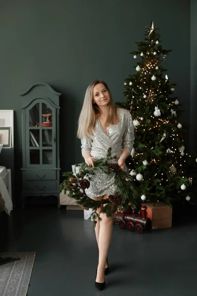 Blond model meisje in een korte modieuze jurk houdt in haar handen een kerstkrans naast een versierde kerstboom — Stockfoto