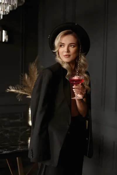 세련 된 검은 옷을 입은 금발의 여자가 호화 로운 실내에서 와인 잔을 들고 포즈를 취한다 — 스톡 사진