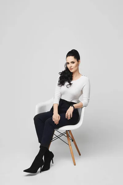 Uzun siyah saçlı, kendine güvenen muhteşem genç kadın gri arka planda sandalyede oturuyor. — Stok fotoğraf