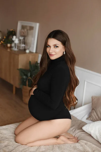 Eine glückliche junge schwangere Frau in einem kuscheligen Strickkleid hält ihre Hände auf ihrem Bauch und blickt in die Kamera — Stockfoto