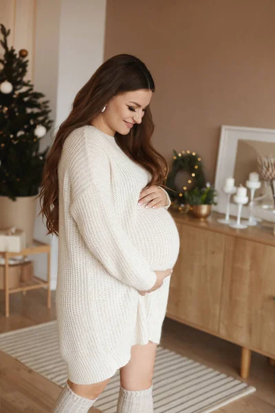 Eine glückliche junge schwangere Frau in einem kuscheligen Strickkleid hält ihre Hände auf ihrem Bauch und betrachtet ihn über das weihnachtlich dekorierte Interieur — Stockfoto